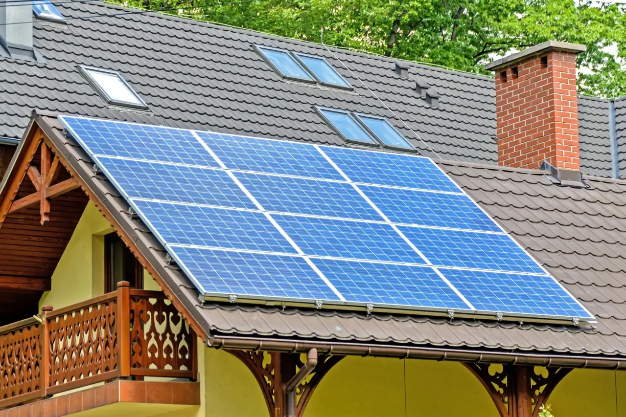 Blaue Solarmodule auf dem Giebeldach eines gelben Hauses mit geschnitztem Holzbalkon (Teising, Landkreis Altötting)