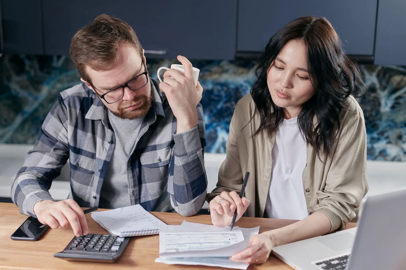 Paar vergleicht am Küchentisch Unterlagen und rechnet mit Taschenrechner und Laptop nach (Ansbach)